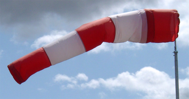 Ein Rot-Weißer Windsack oder Windschlauch wird mit der von rechts nach links strömenden Luft gefüllt und steht so fast senkrecht zur Stabaufstellung, was ein Zeichen für einen windigen Tag ist.