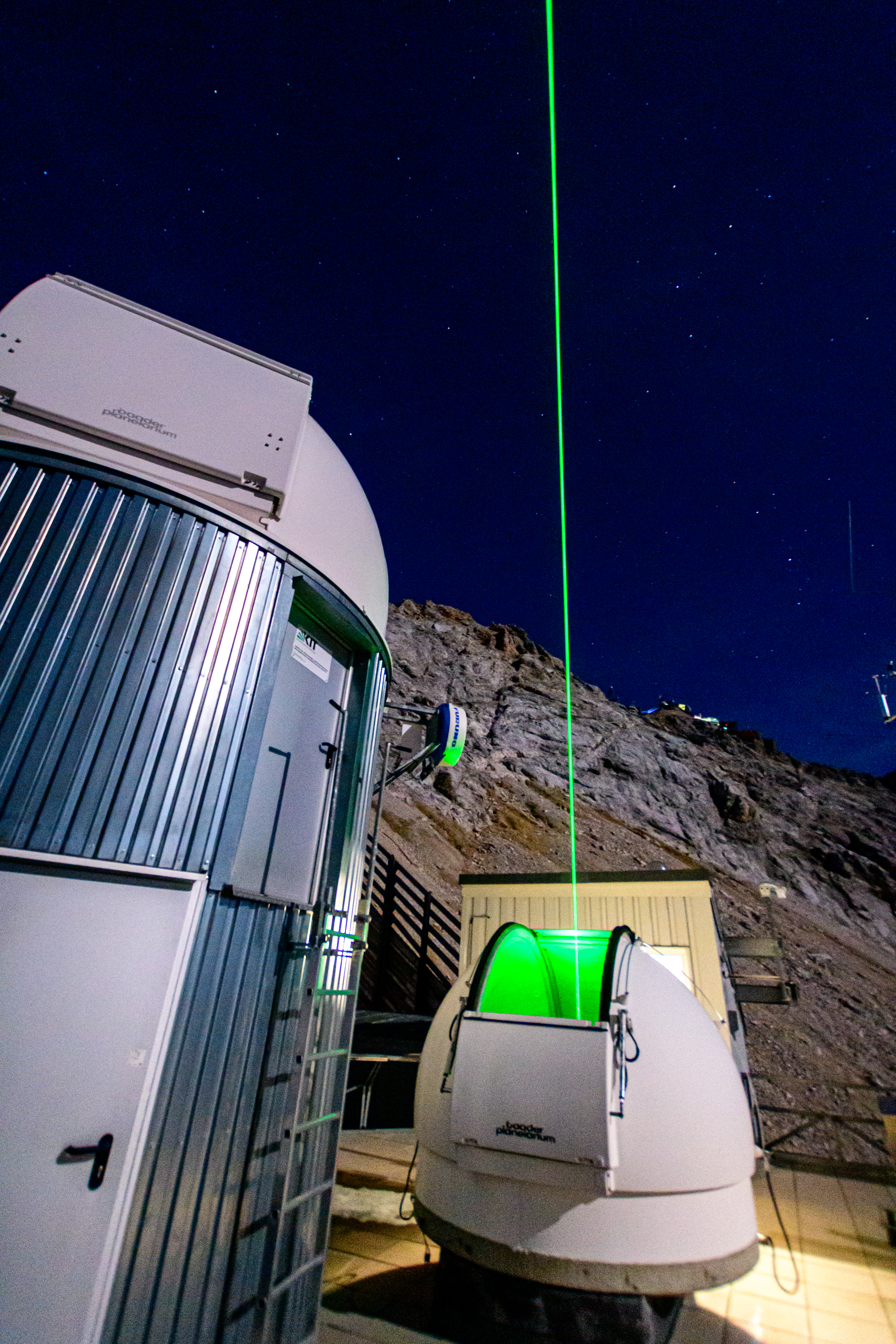 Dieses Bild zeigt wie ein grüner Laserstrahl aus einer alpinen LiDAR-Kuppel in den Nachthimmel vertikal nach oben leuchtet.