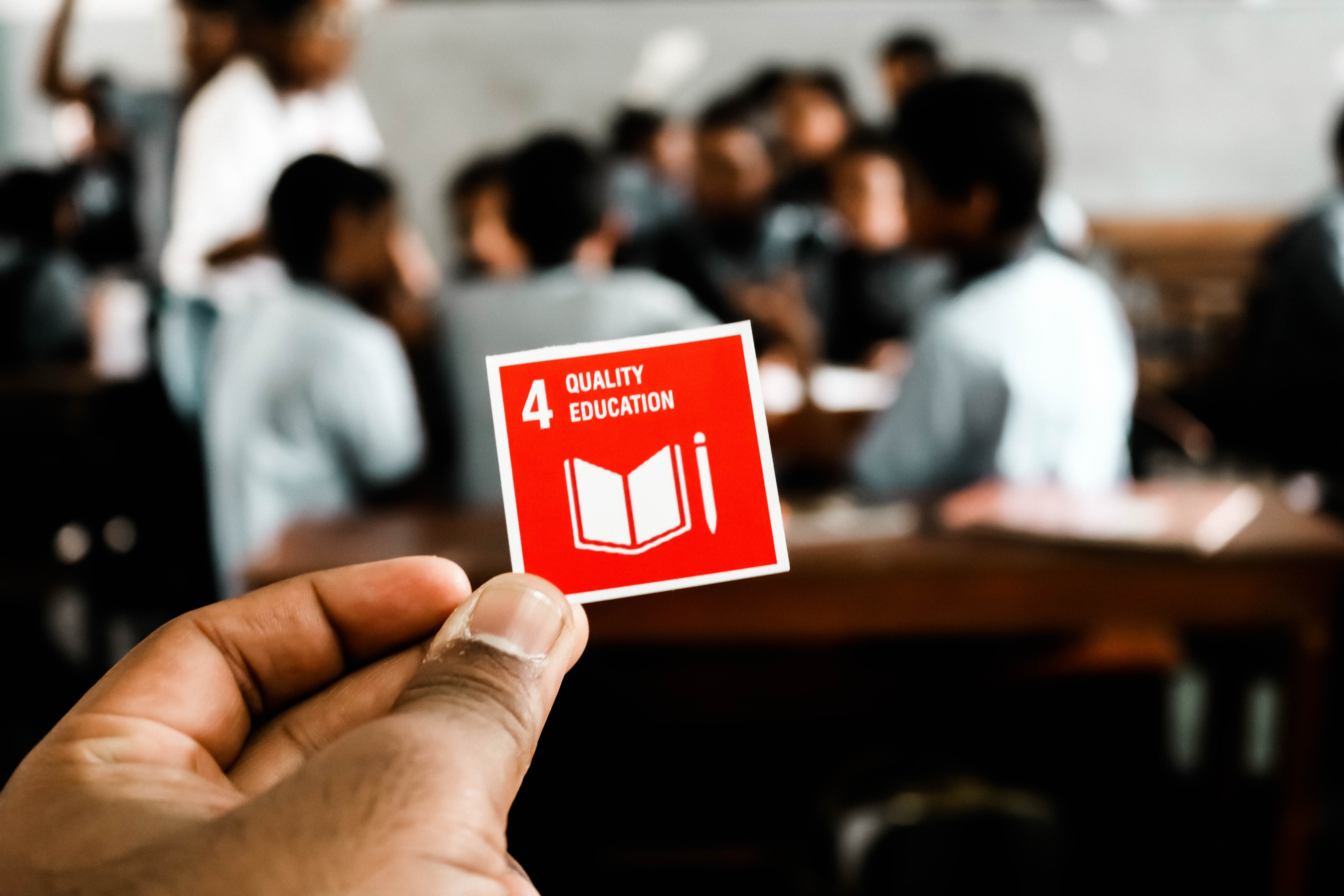 Eine Hand hält das SDG 4 Logo im Vordergrund. Dahinter sind sehr unscharf Schüler*innen an Tischen zu sehen.